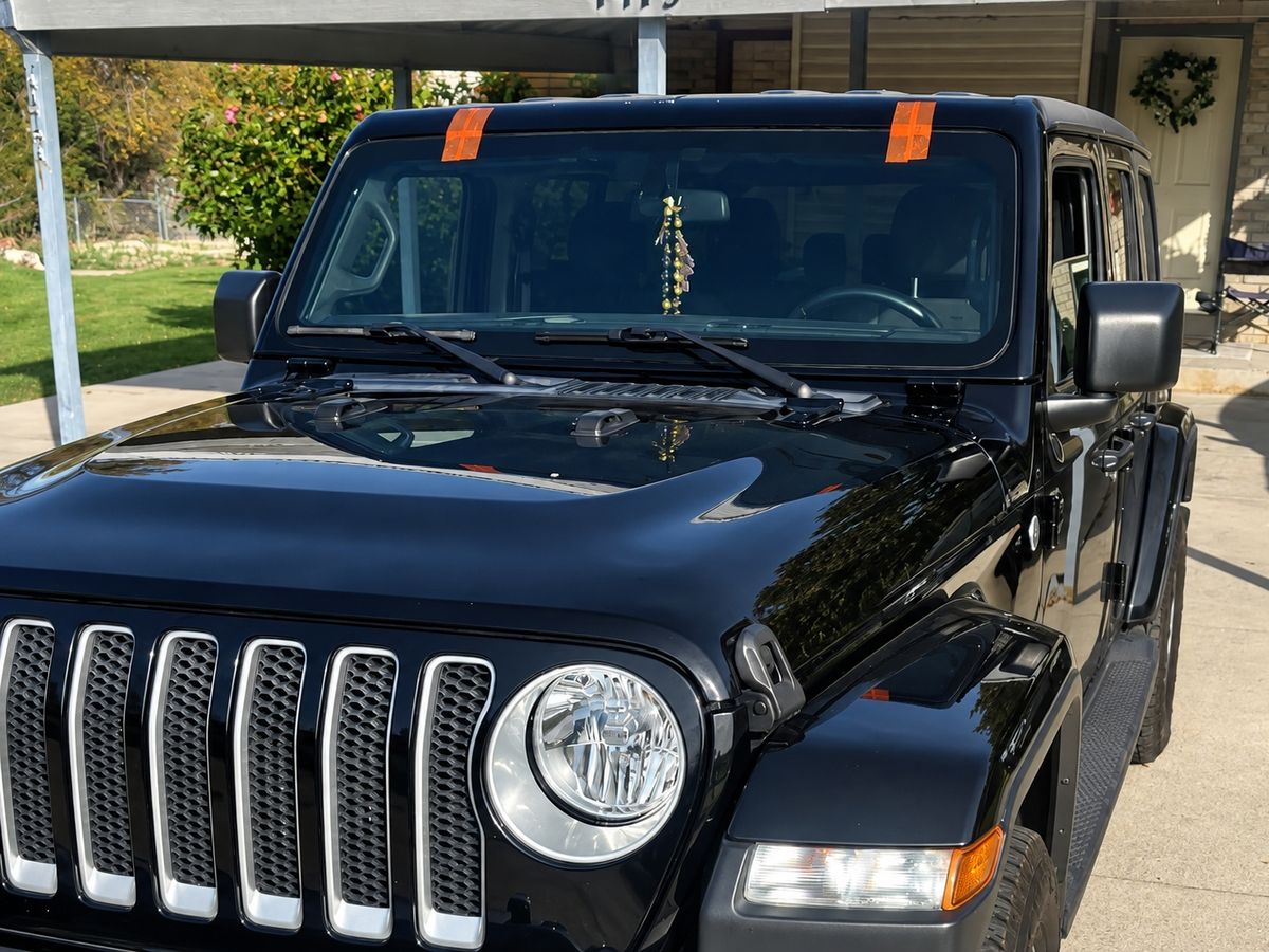 Black four-door Jeep Wrangler under a carport, its new windshield taped at the top